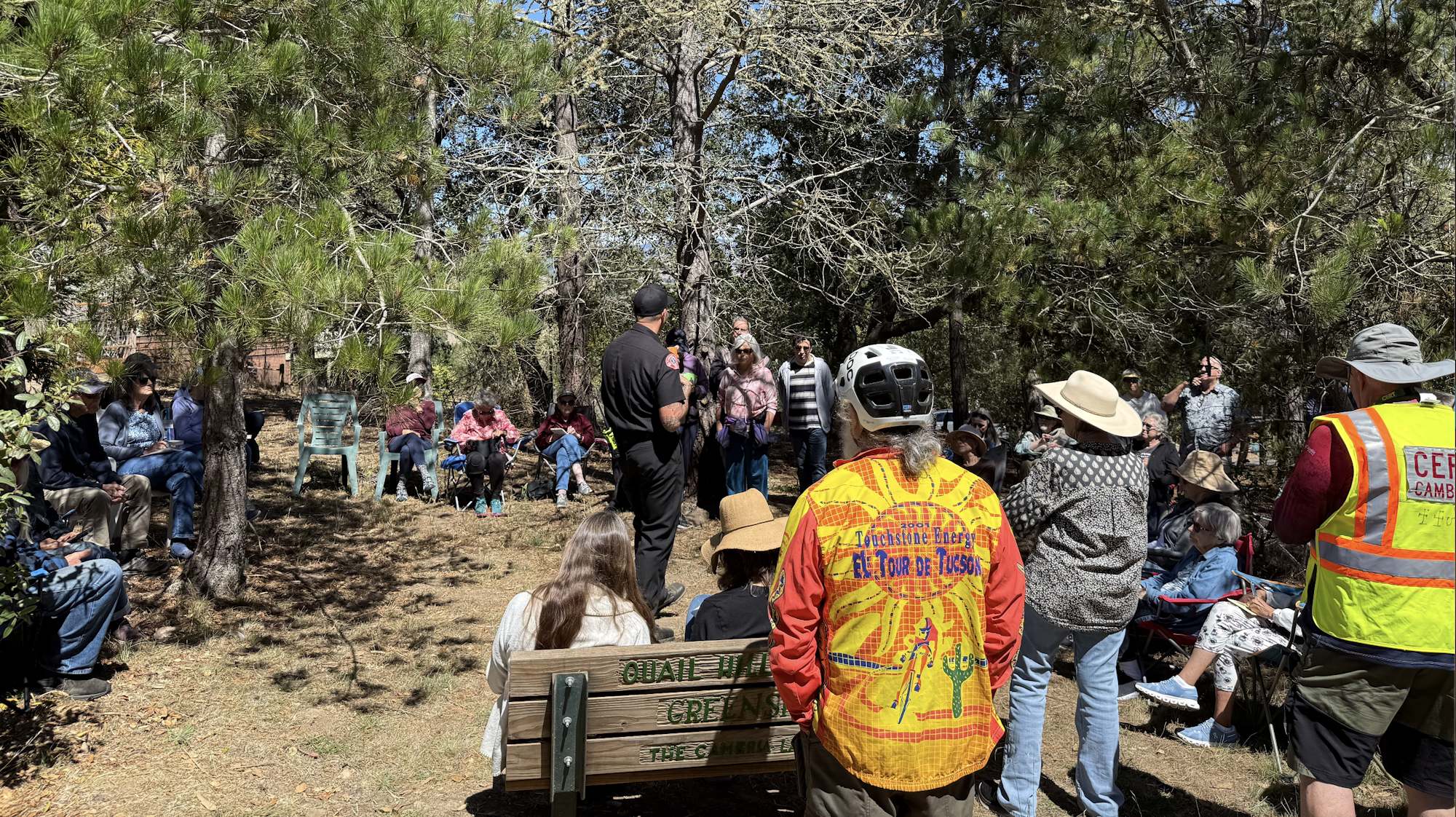 A gathering of people outdoors, mostly seated, listening to a speaker. Some are wearing sun hats and bright vests. Trees in the background.