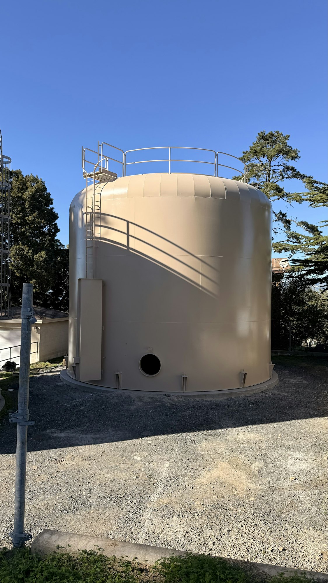 The image shows a large cylindrical tank, likely for storing liquids, with stairs and a clear blue sky in the background.