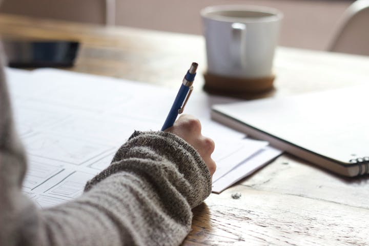 Person writing on paper, coffee cup in background on wooden table.