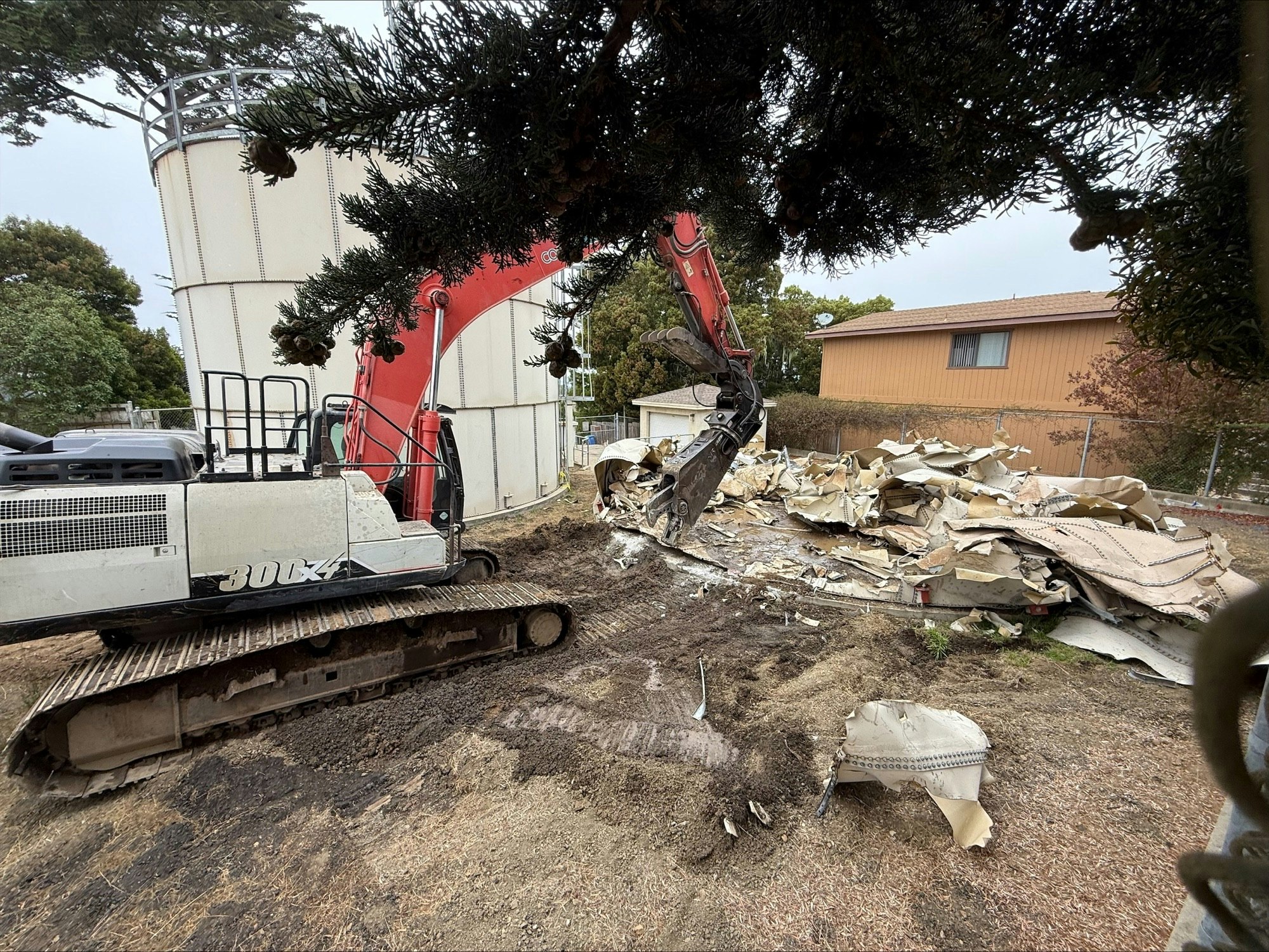 Excavator demolishing a structure, debris piled nearby, with trees and a building in the background.
