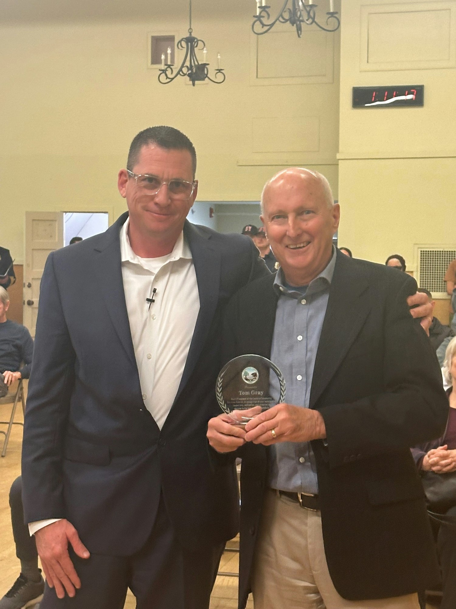 Two men smiling, one holding an award, in a room with people seated in the background.