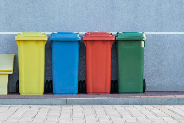 Four colorful wheelie bins (yellow, blue, red, green) lined up against a gray wall on pavement.