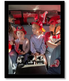 Children and a firefighter in a vehicle, kids wearing red fire helmets.