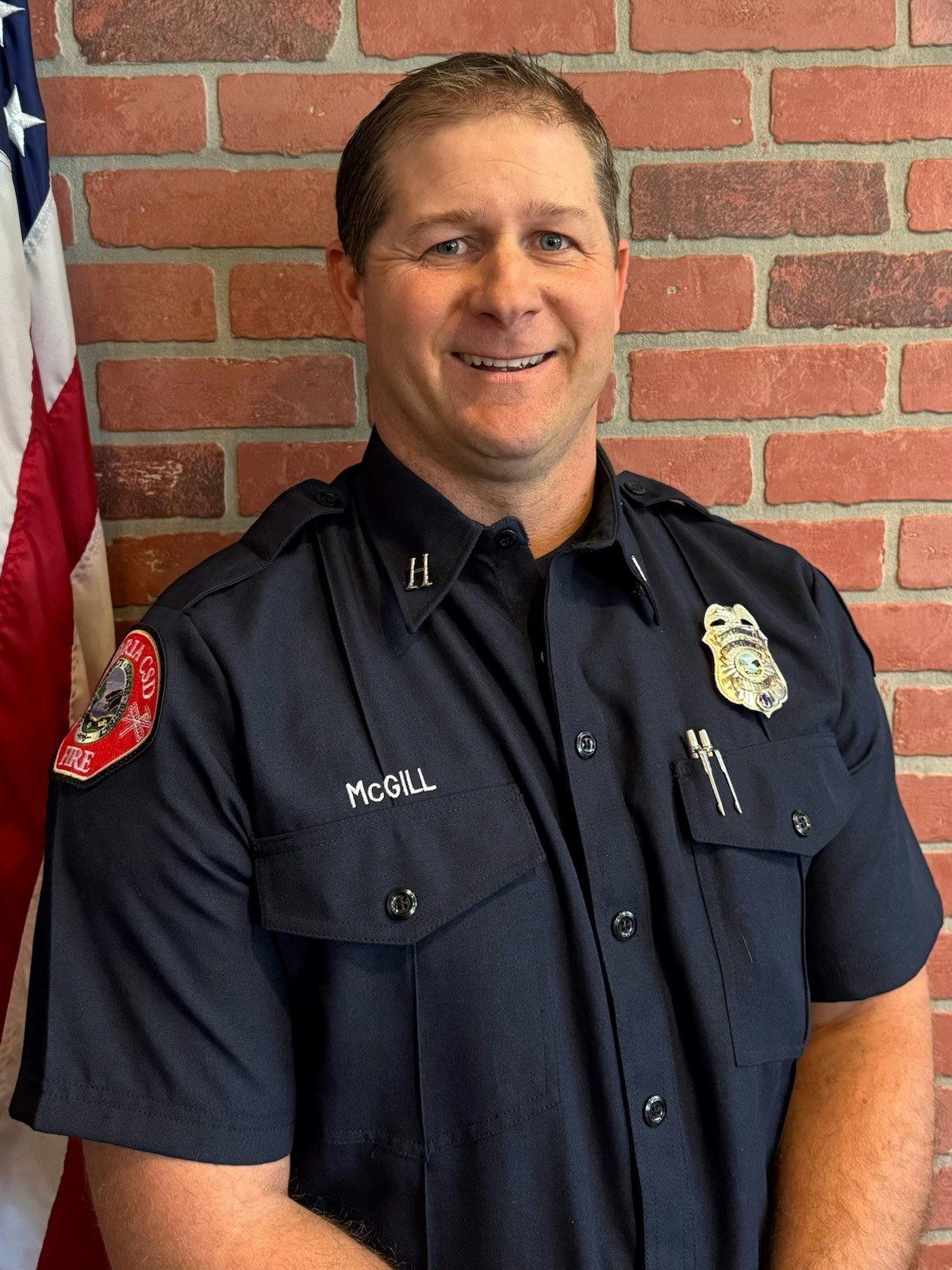 A person in a uniform with a badge, standing in front of a brick wall and an American flag.