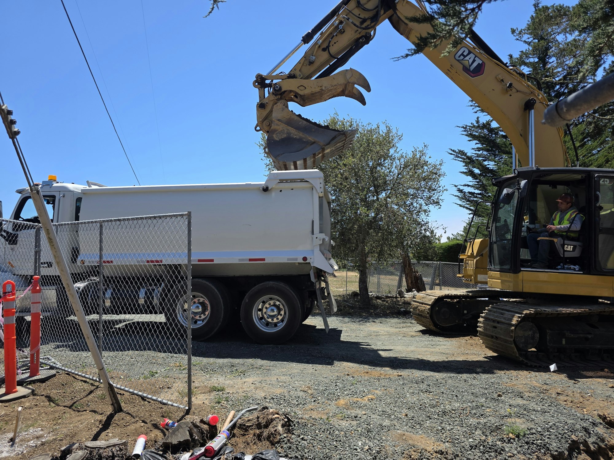 A construction site with a dump truck and an excavator, where a worker operates the machinery under a clear blue sky.