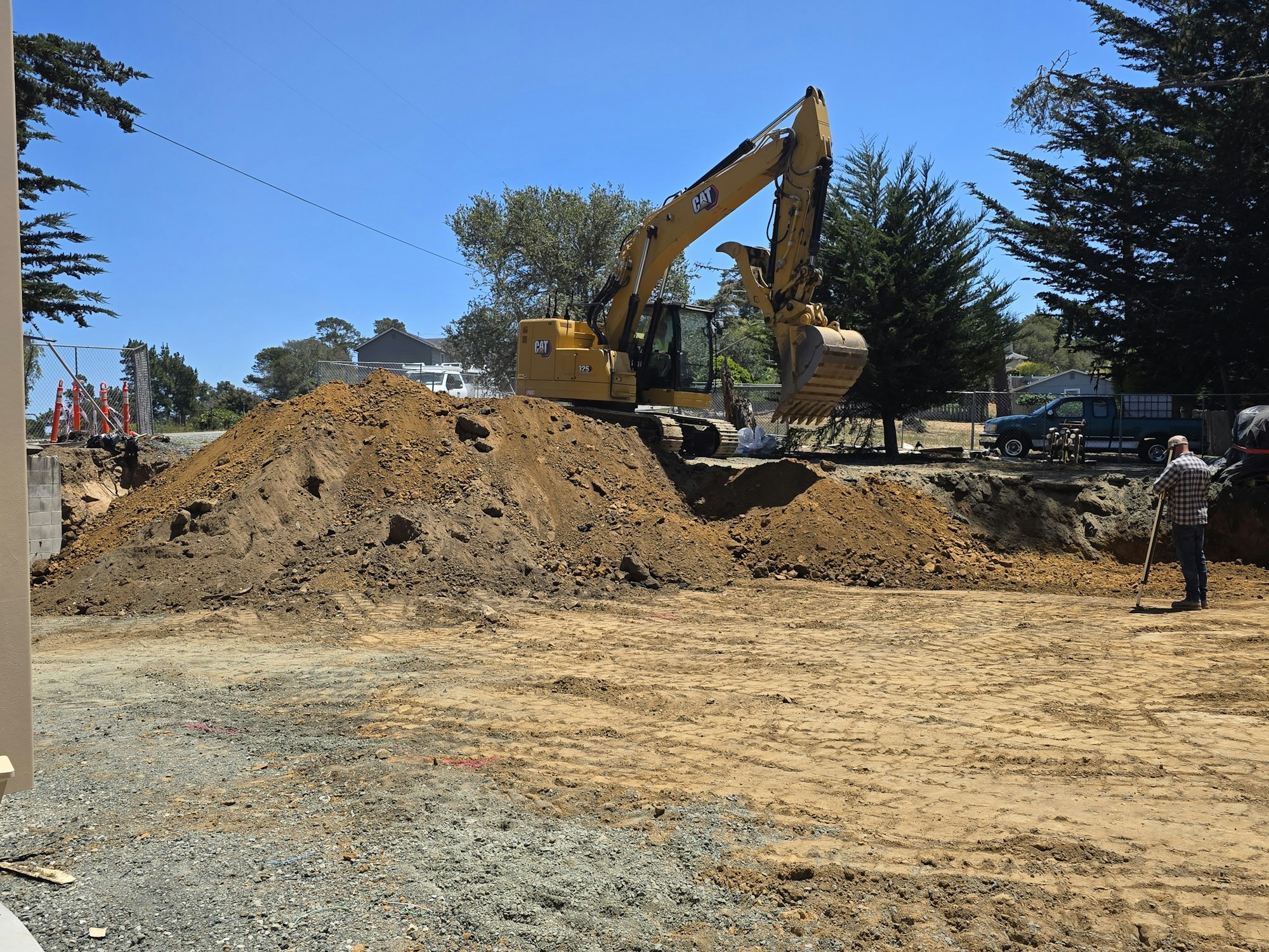 A construction site with a large excavator, a dirt mound, and a person working nearby under a clear blue sky.