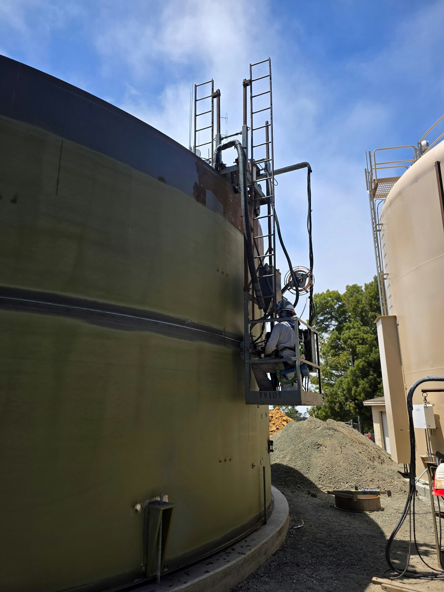 A worker on a lift is inspecting or maintaining a large tank, surrounded by another tank and piles of gravel.