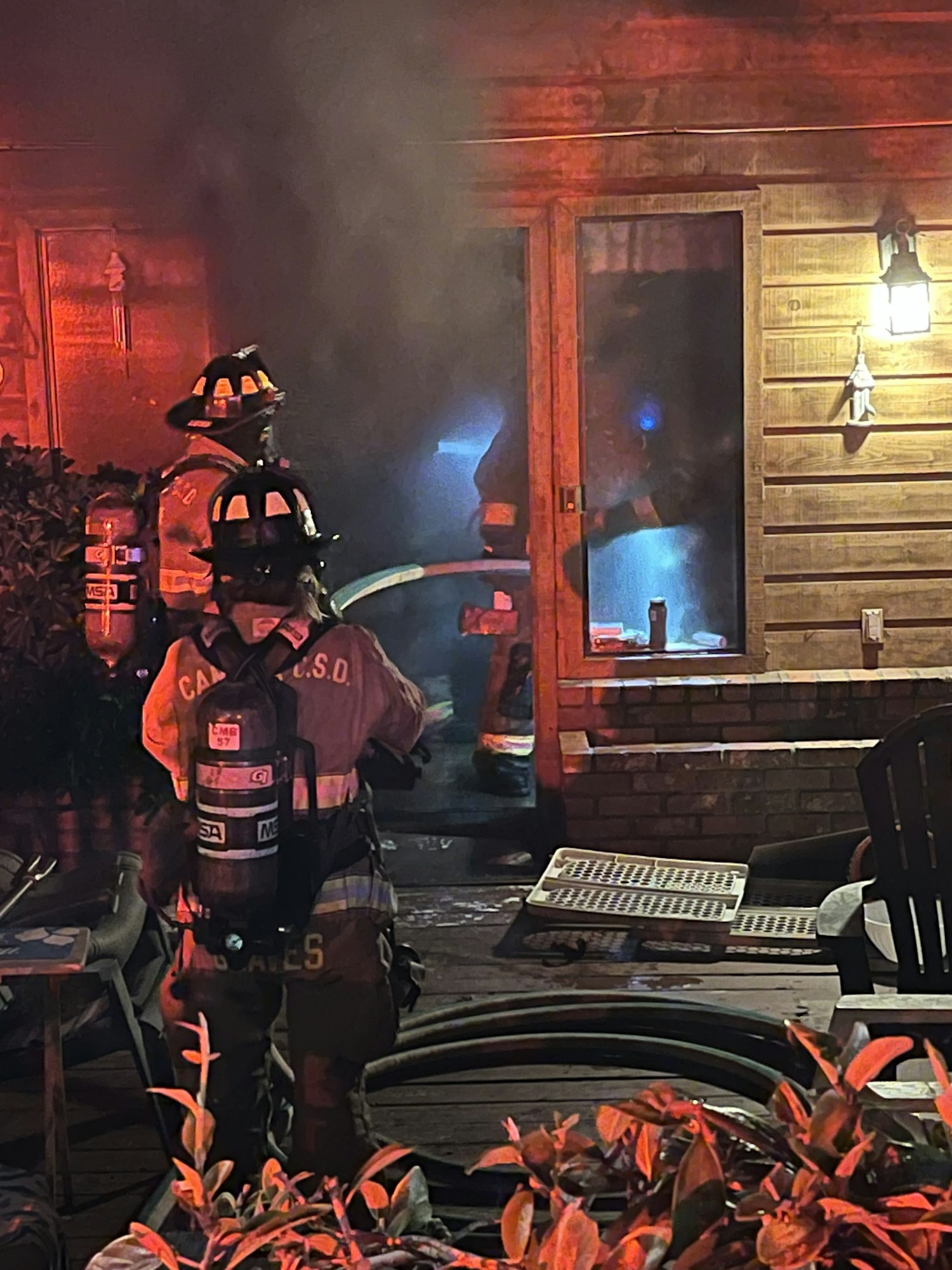 Firefighters in gear, managing a smoky scene at night by a wooden structure with a hose.