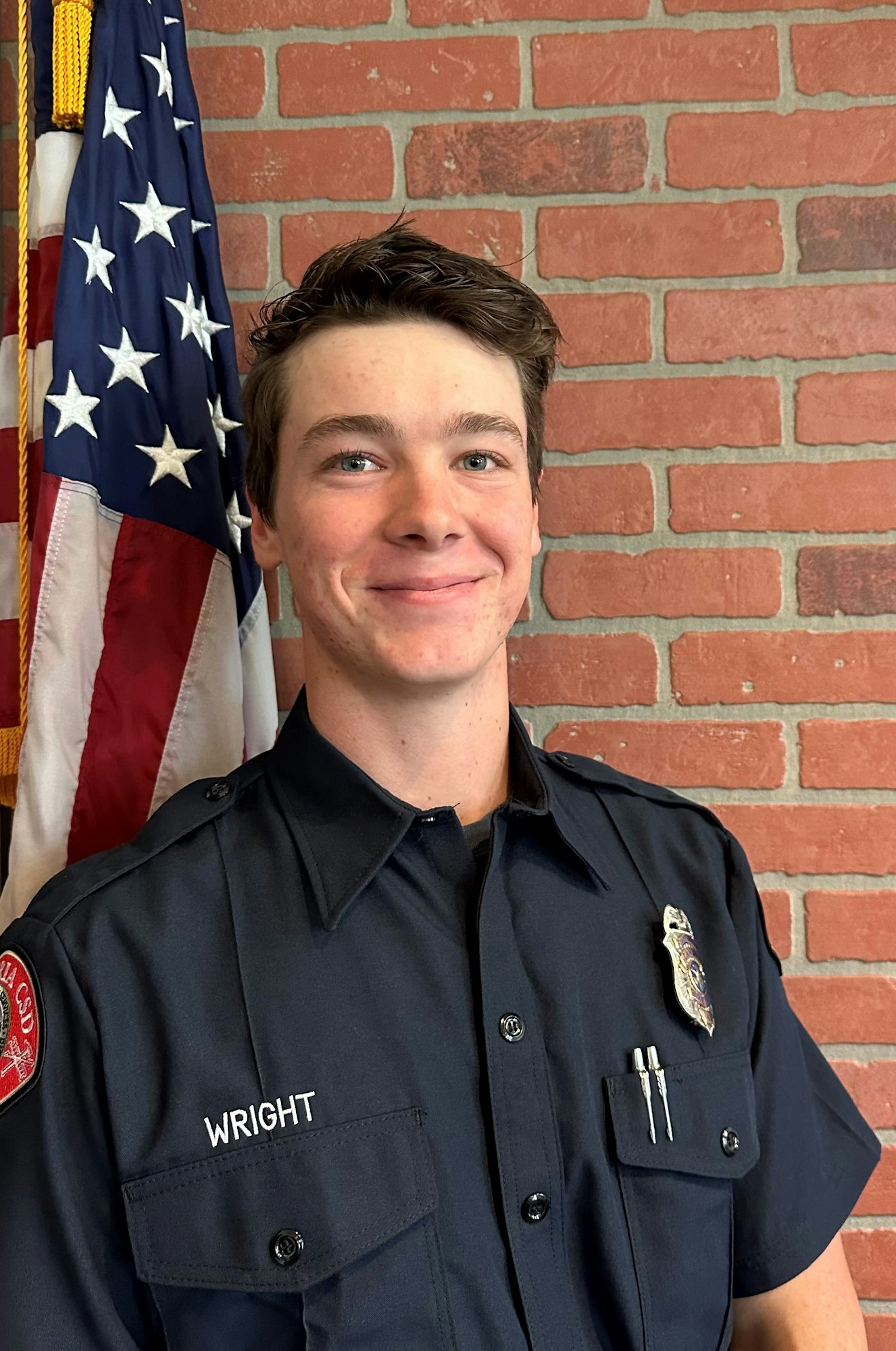 A young man in a blue uniform with the name "Wright" stands next to an American flag, smiling against a brick wall.