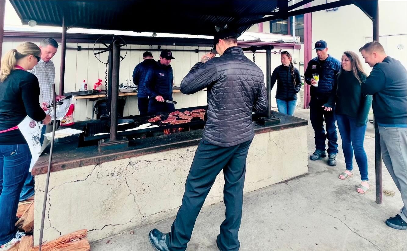 Group of people gathered around an outdoor grill with some cooking meat.