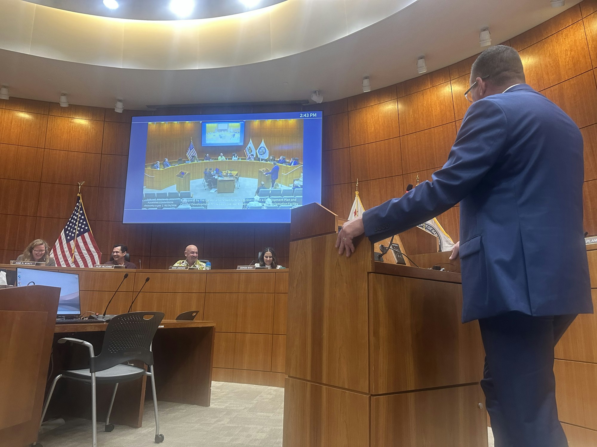 A formal meeting in a chamber, featuring a speaker at a podium, officials seated, and a large screen displaying participants.