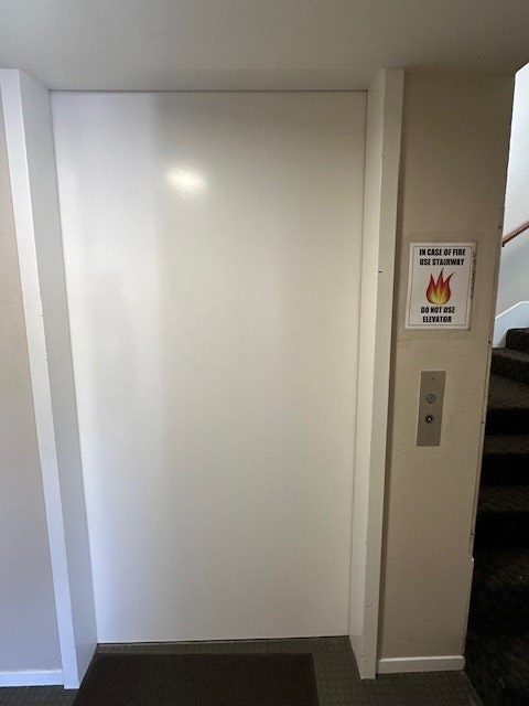 An elevator door with a fire safety instruction sign and a call button, next to a carpeted stairway.