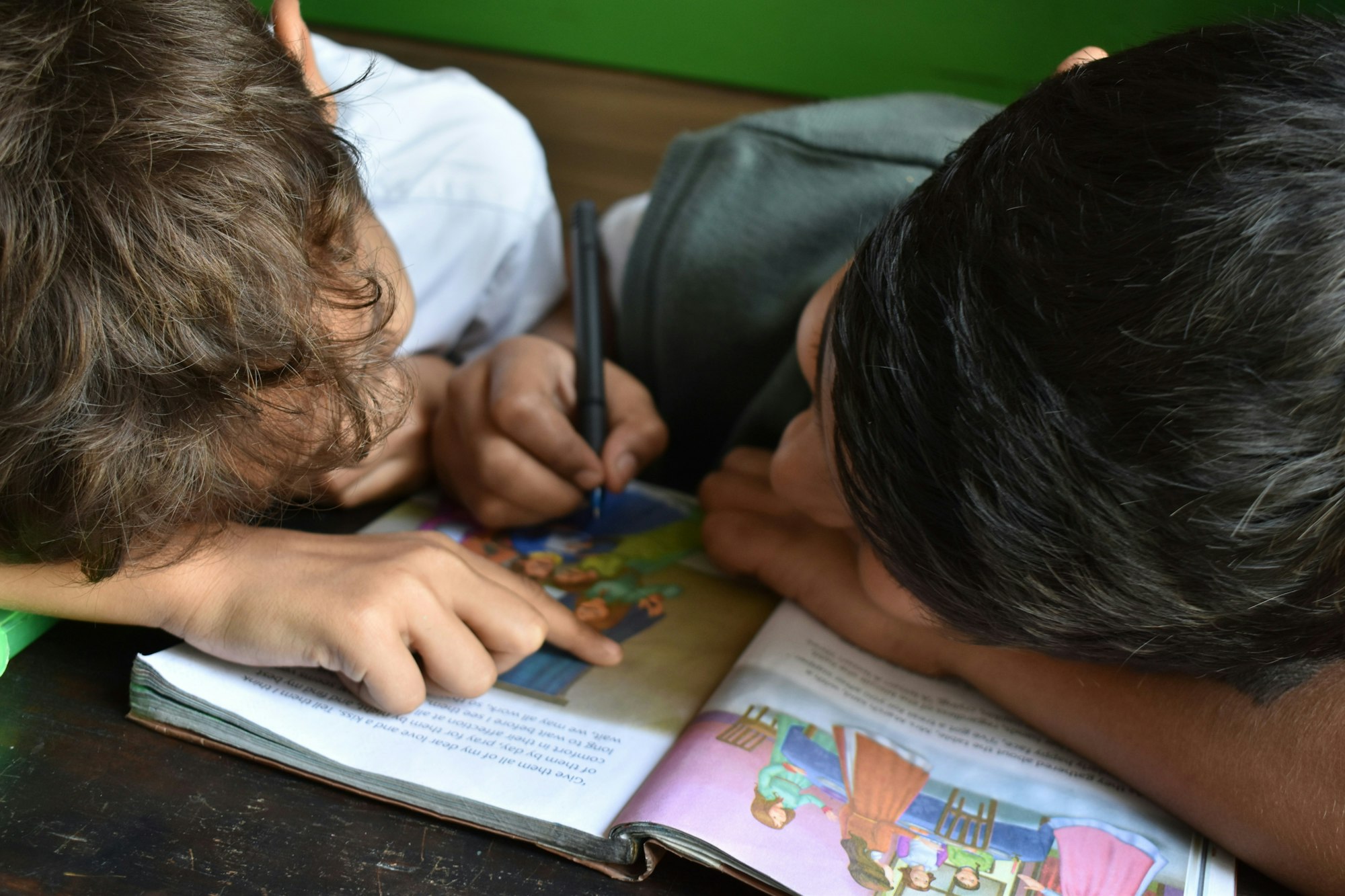 Two children reading a book together, one is pointing while the other writes.
