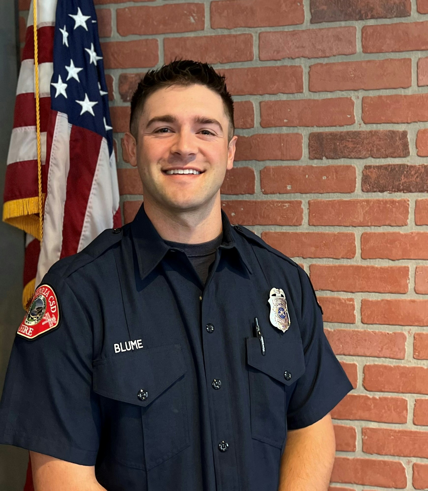 A smiling person in a fire department uniform stands in front of a brick wall and an American flag.