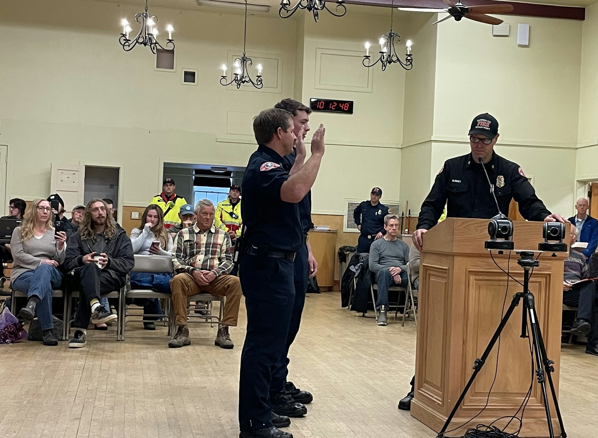 A firefighter is being sworn in at a ceremony, with an audience seated and others standing around.