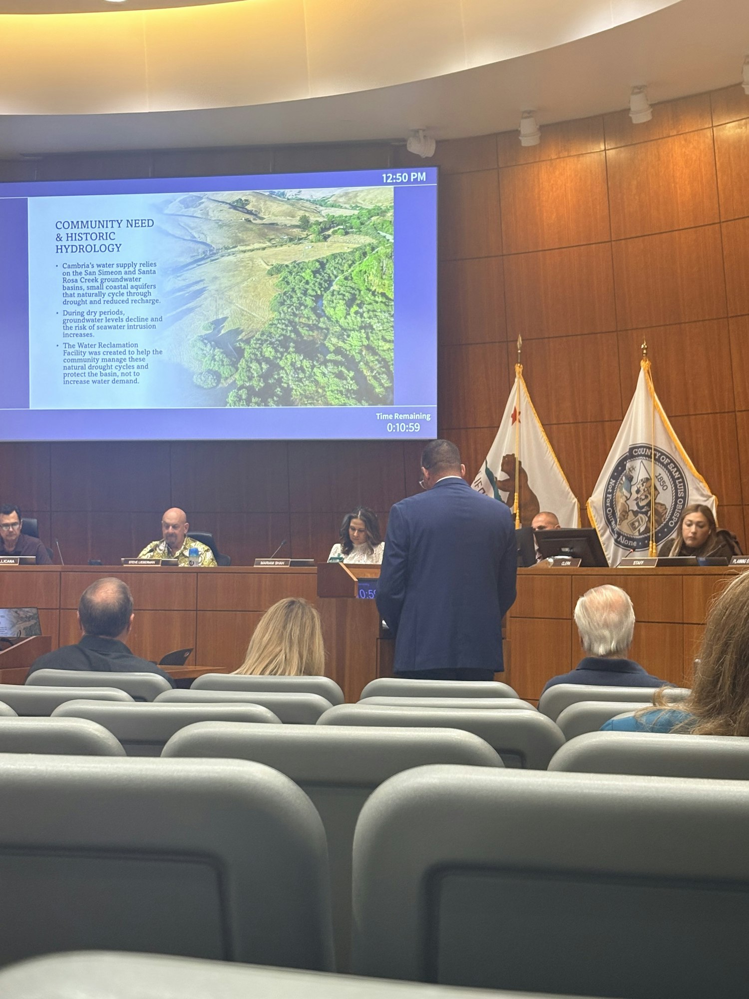 A meeting is taking place with a slide presentation discussing community needs and historic hydrology, shown on a screen.