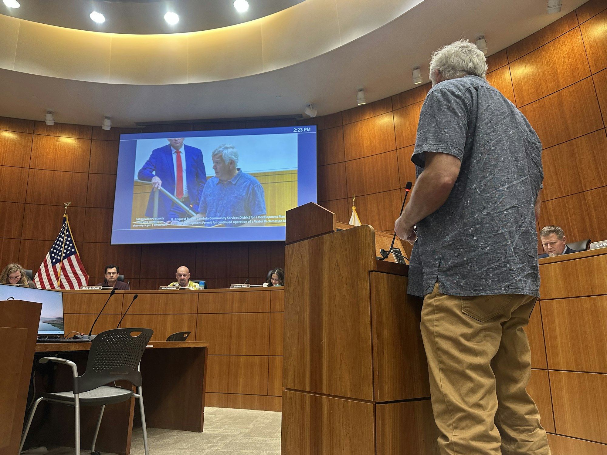 A person is speaking at a public meeting, with officials seated at a panel and a screen displaying visuals in the background.
