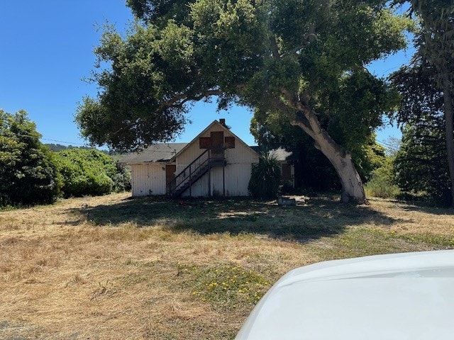 A weathered barn sits behind a dry field, shaded by a large tree under a clear blue sky. There's a car's hood at the bottom edge.