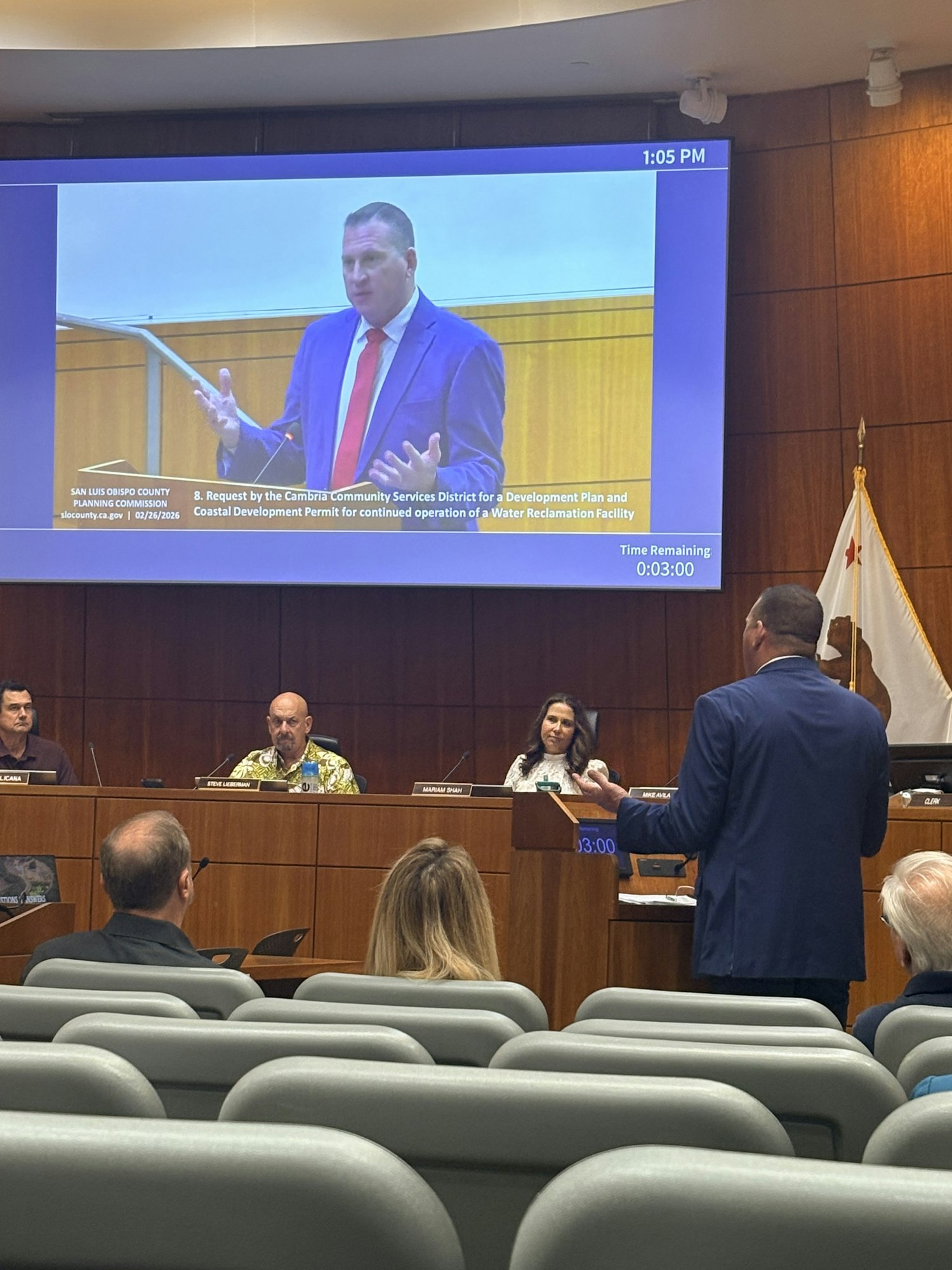 A meeting or hearing in progress, with a speaker presenting on a screen and an audience seated behind.