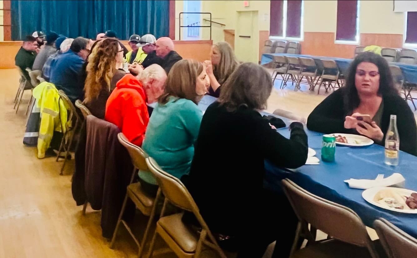 People seated at tables in a hall, some eating, one person looking at a smartphone.