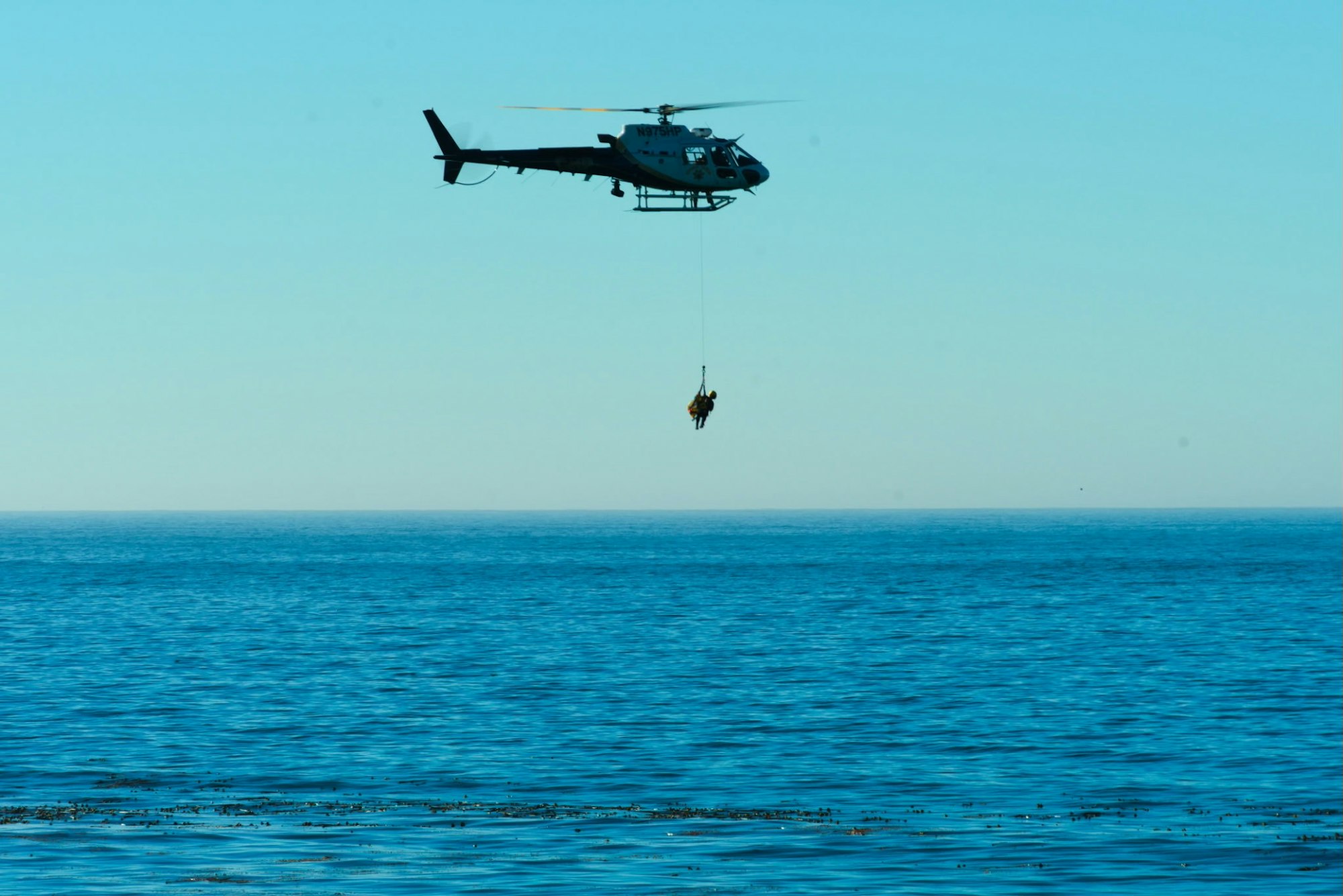 A helicopter hovers above the ocean, lowering a person using a rescue line against a clear blue sky.