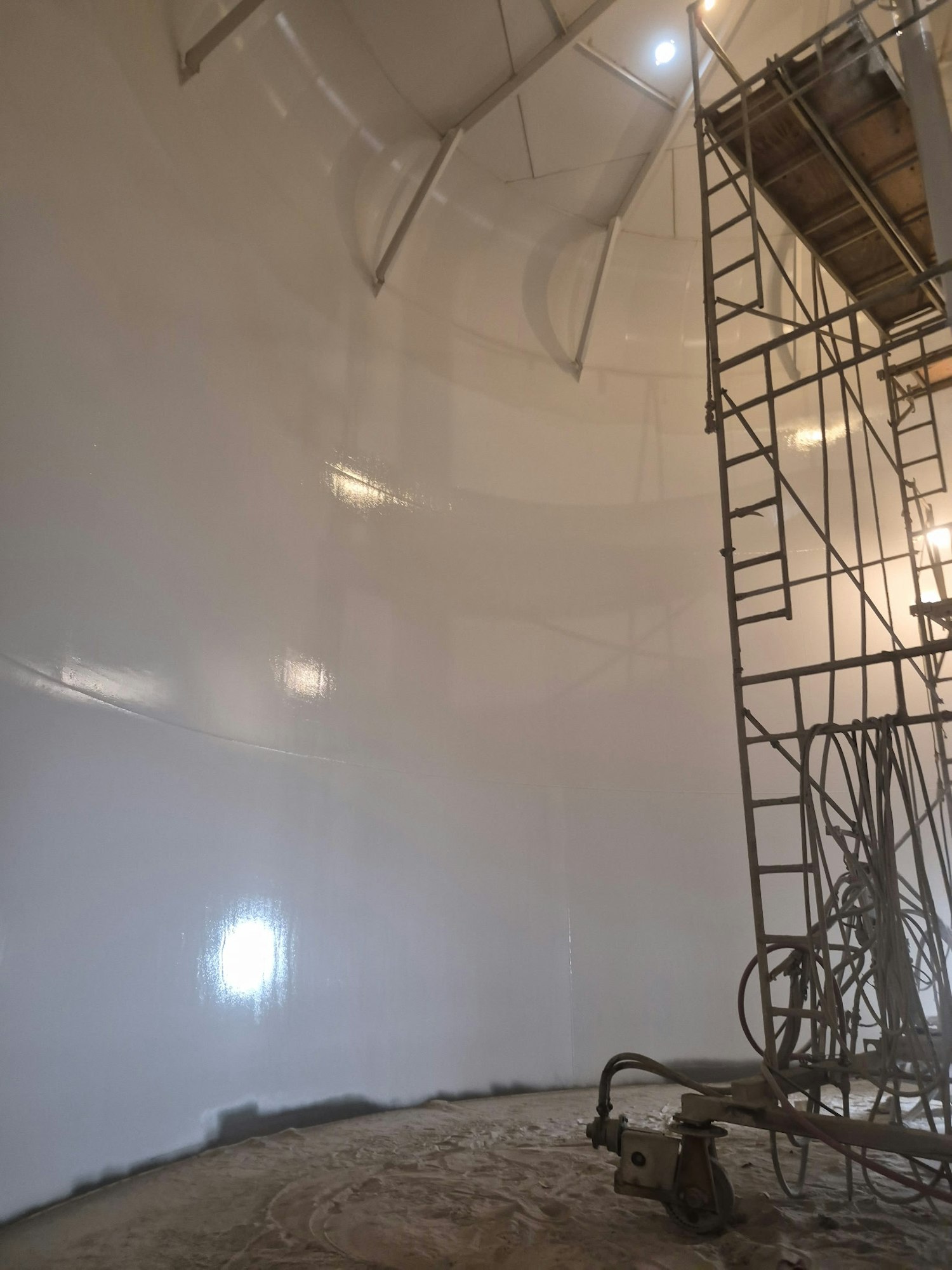 The image shows a large, white, cylindrical space with a metal scaffolding and equipment for construction or painting work.
