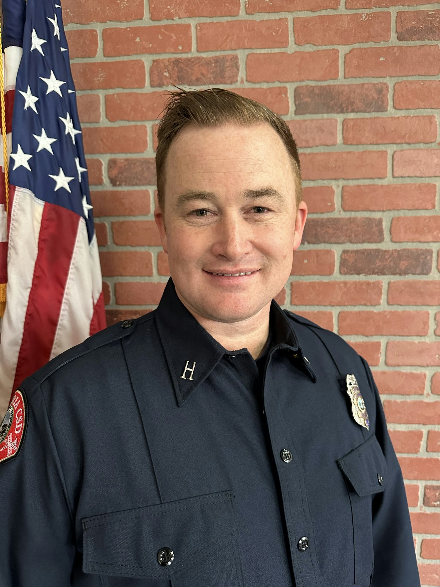A person in uniform with a badge, tilting head, against a US flag and brick wall background. (The image is rotated)