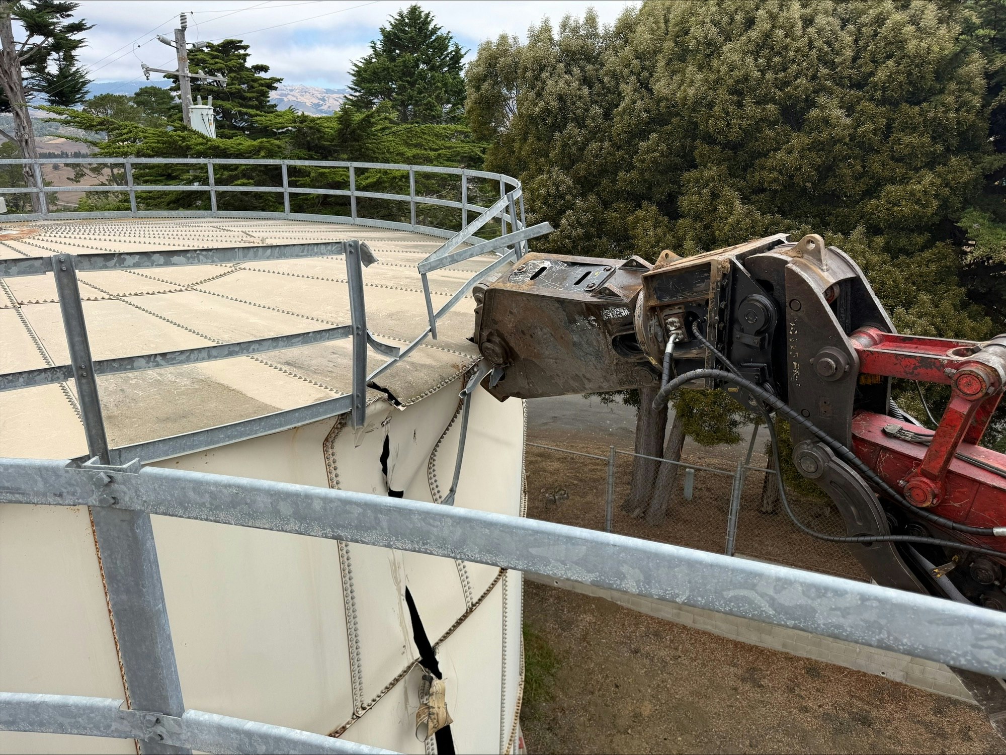 A mechanical claw is seen tearing through a metal storage tank beside a safety rail.