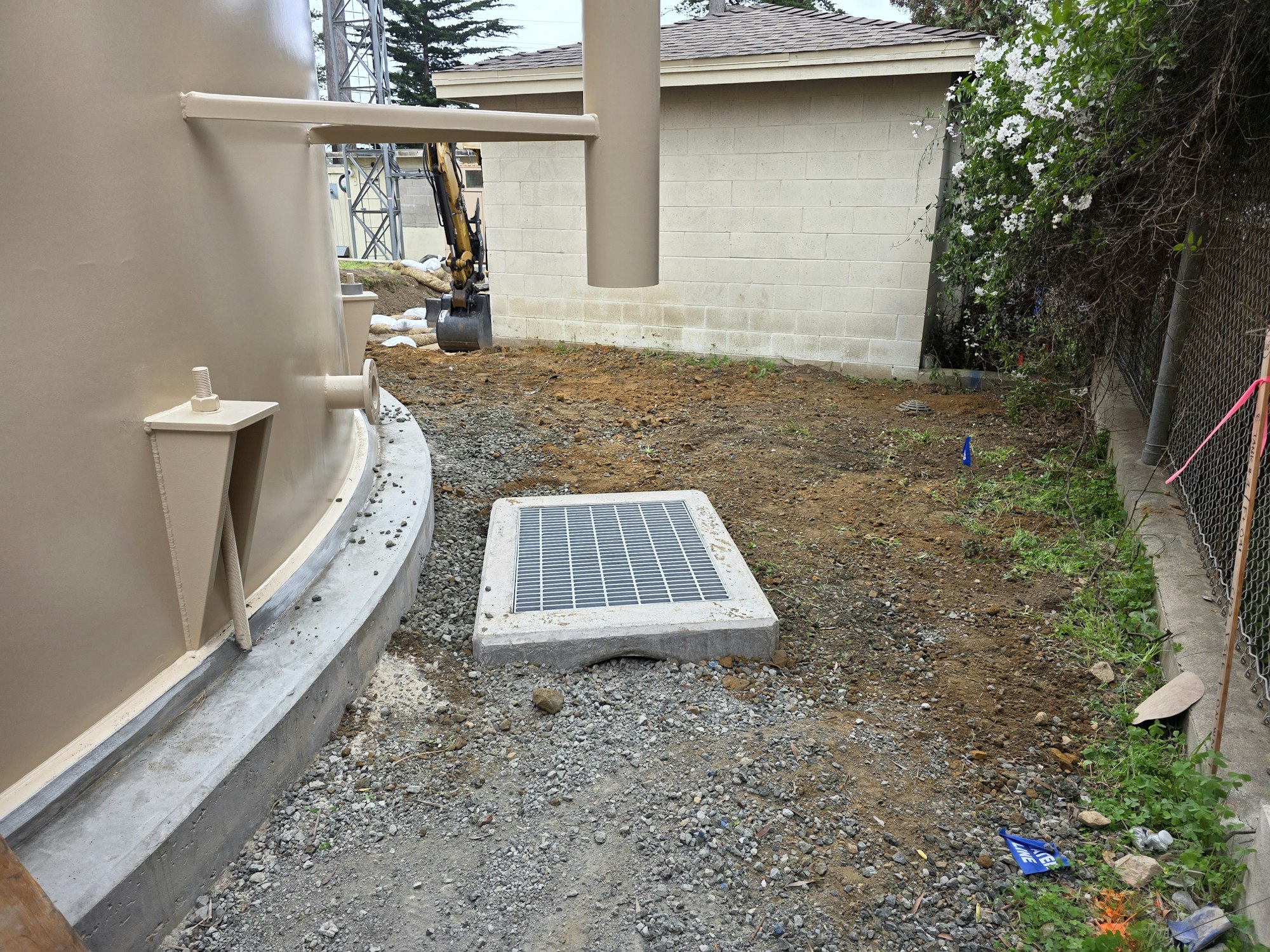 The image shows a construction area with a large tan tank, a drainage grate, gravel, and a nearby building and fence.