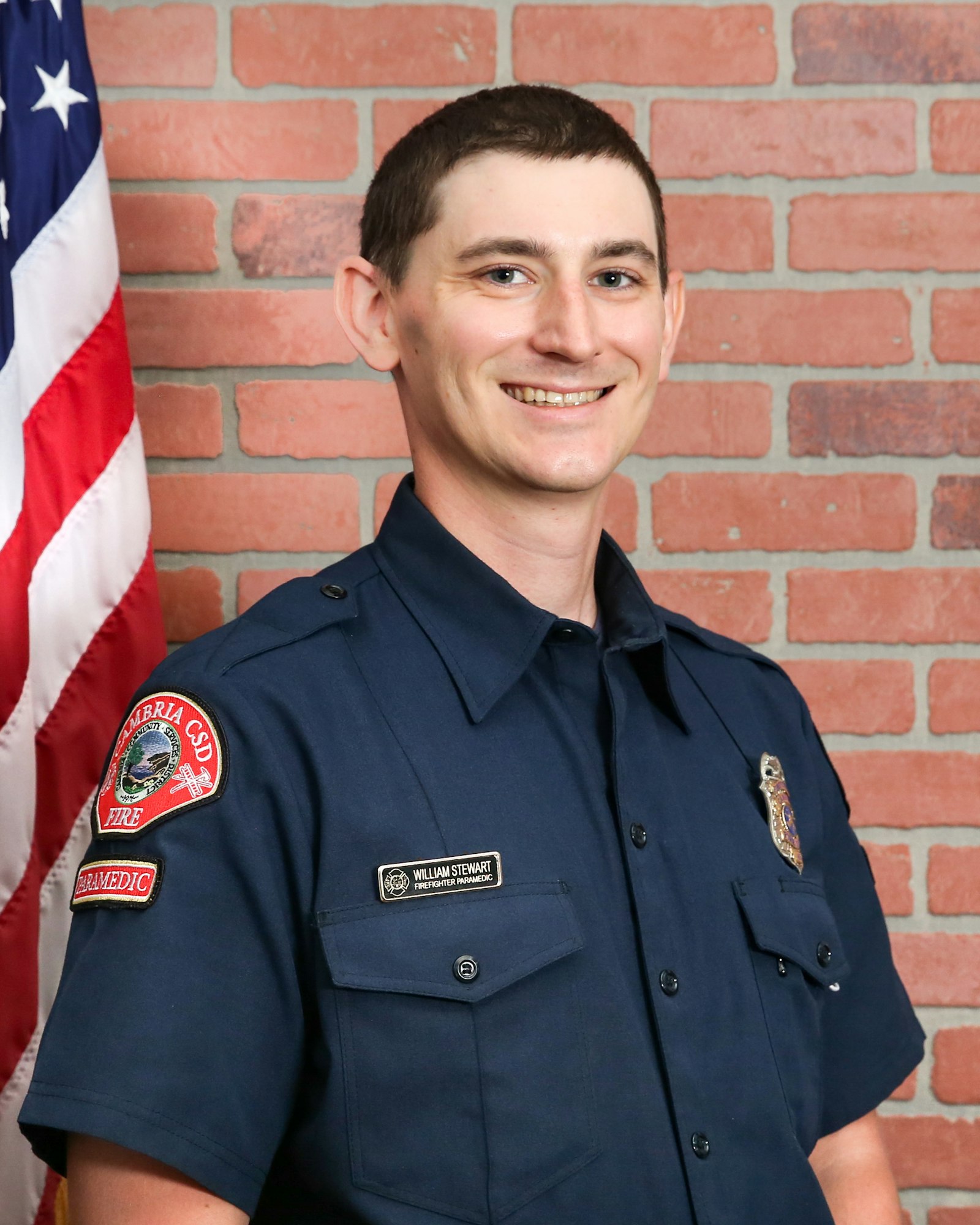 A smiling man in a dark blue uniform stands in front of a brick wall and an American flag, representing a fire paramedic role.