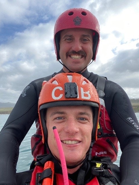 Two men in helmets are smiling while kayaking on a body of water, dressed in wet suits, with a cloudy sky above.