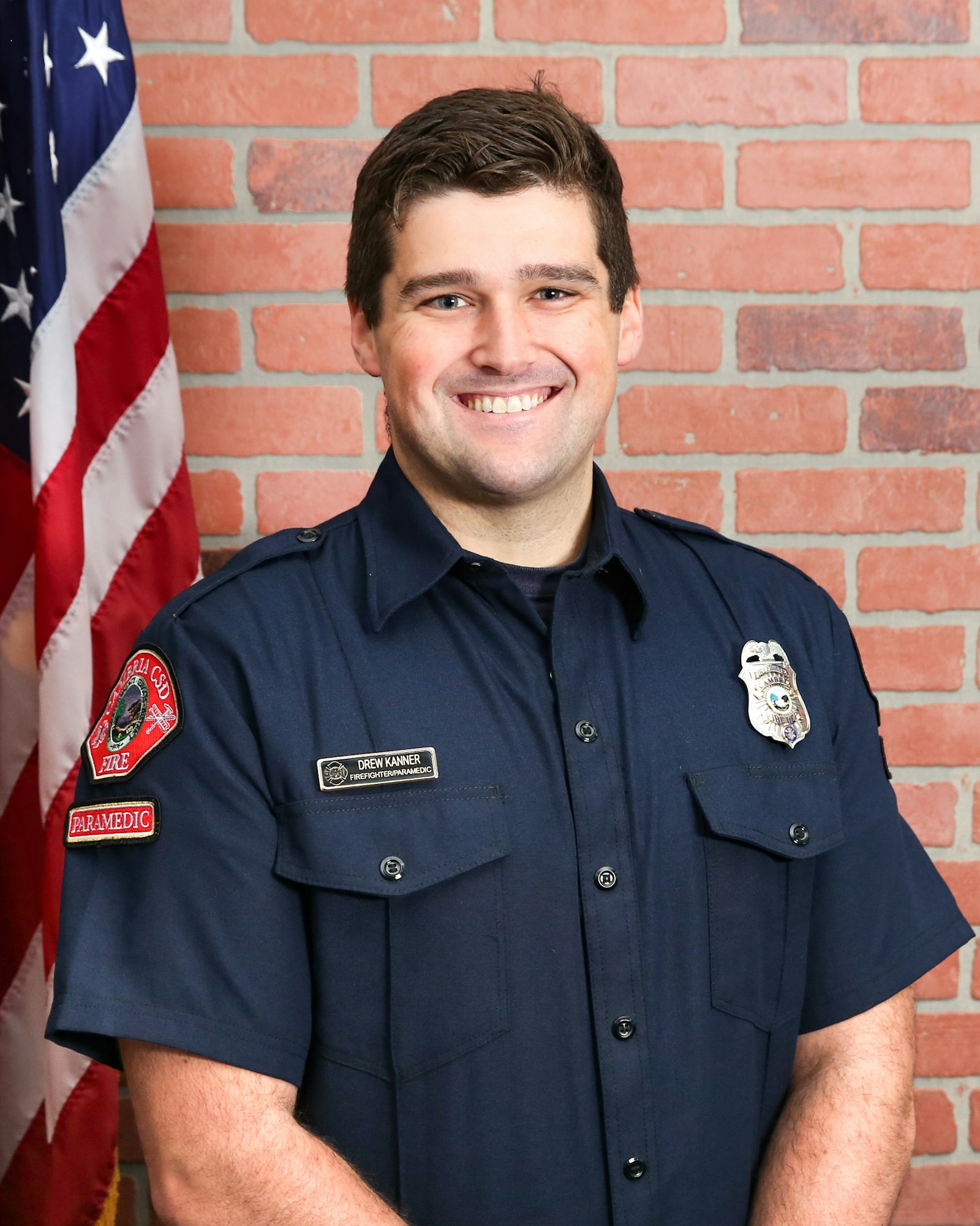 A smiling firefighter in uniform stands in front of a brick wall and an American flag.