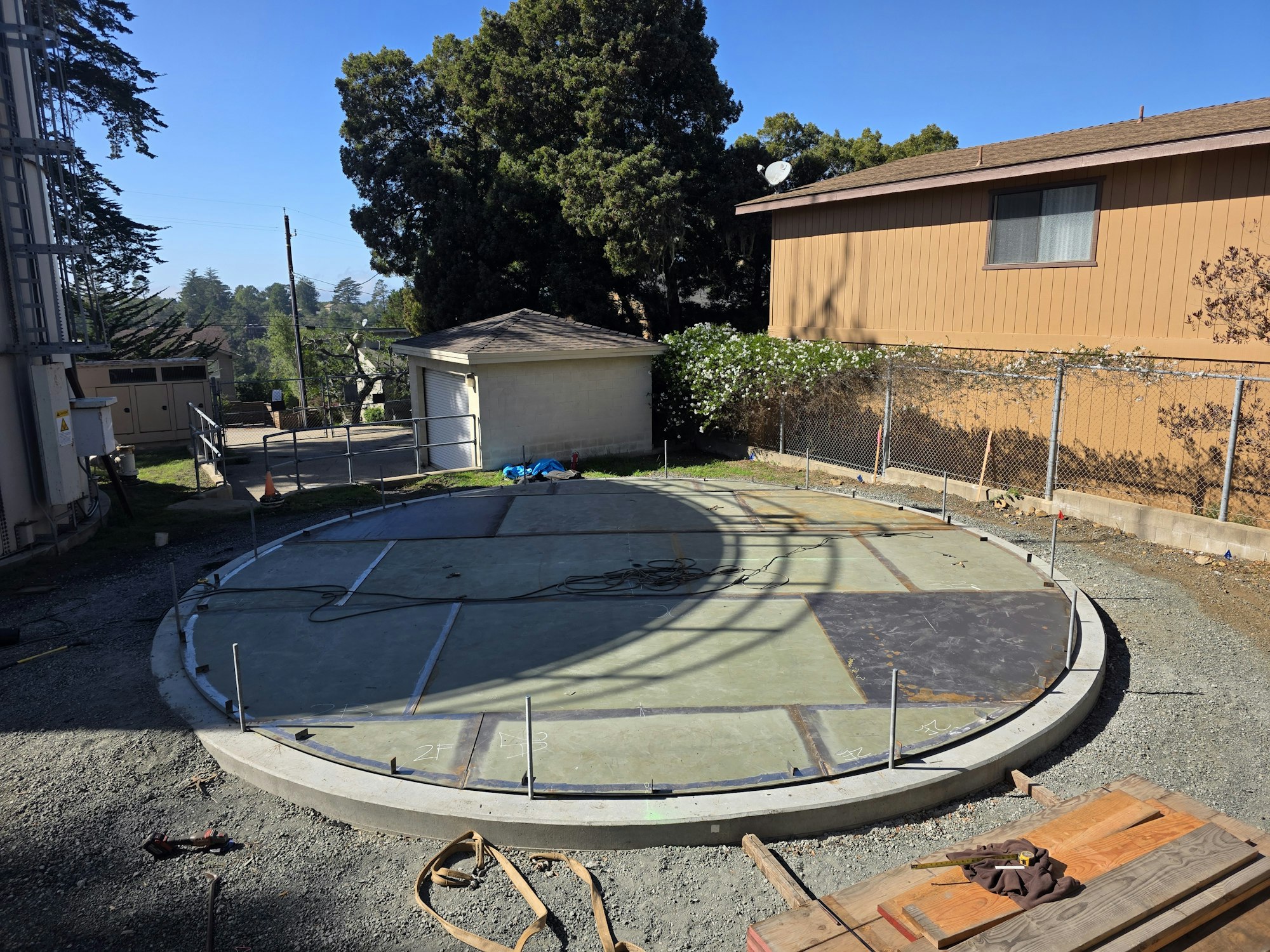 A circular concrete pad in a yard with nearby trees, a building, and scattered tools.