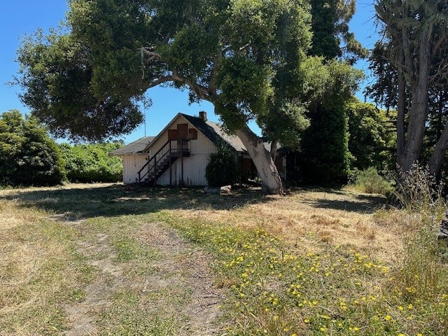 An old house with a staircase outside, surrounded by trees and overgrown grass under a clear sky.