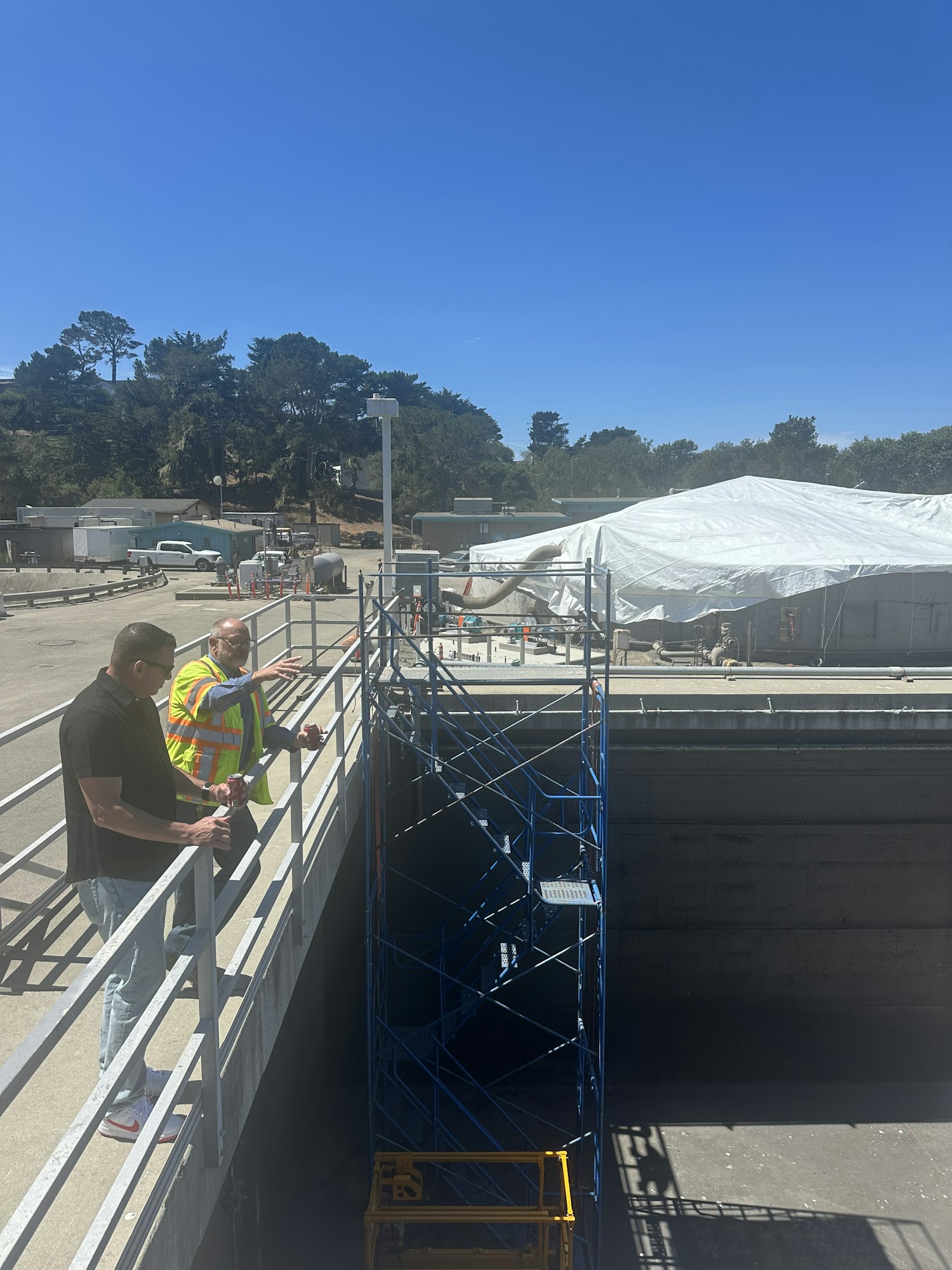 Two people in safety gear near a large scaffolding and covered structure on a sunny day.