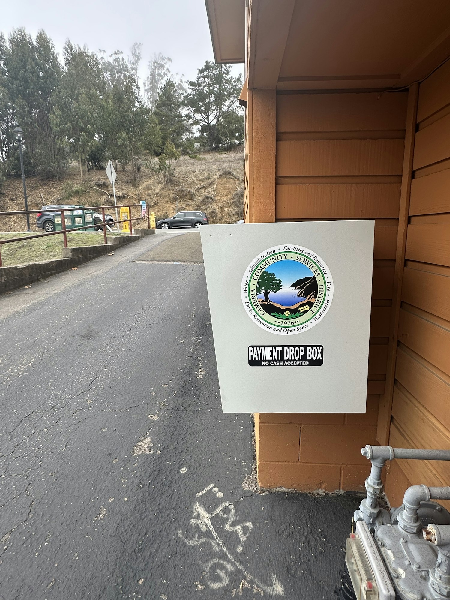 A payment drop box is attached to a wall, featuring a logo and text "No cash accepted." It's outdoors, with trees and cars in the background.