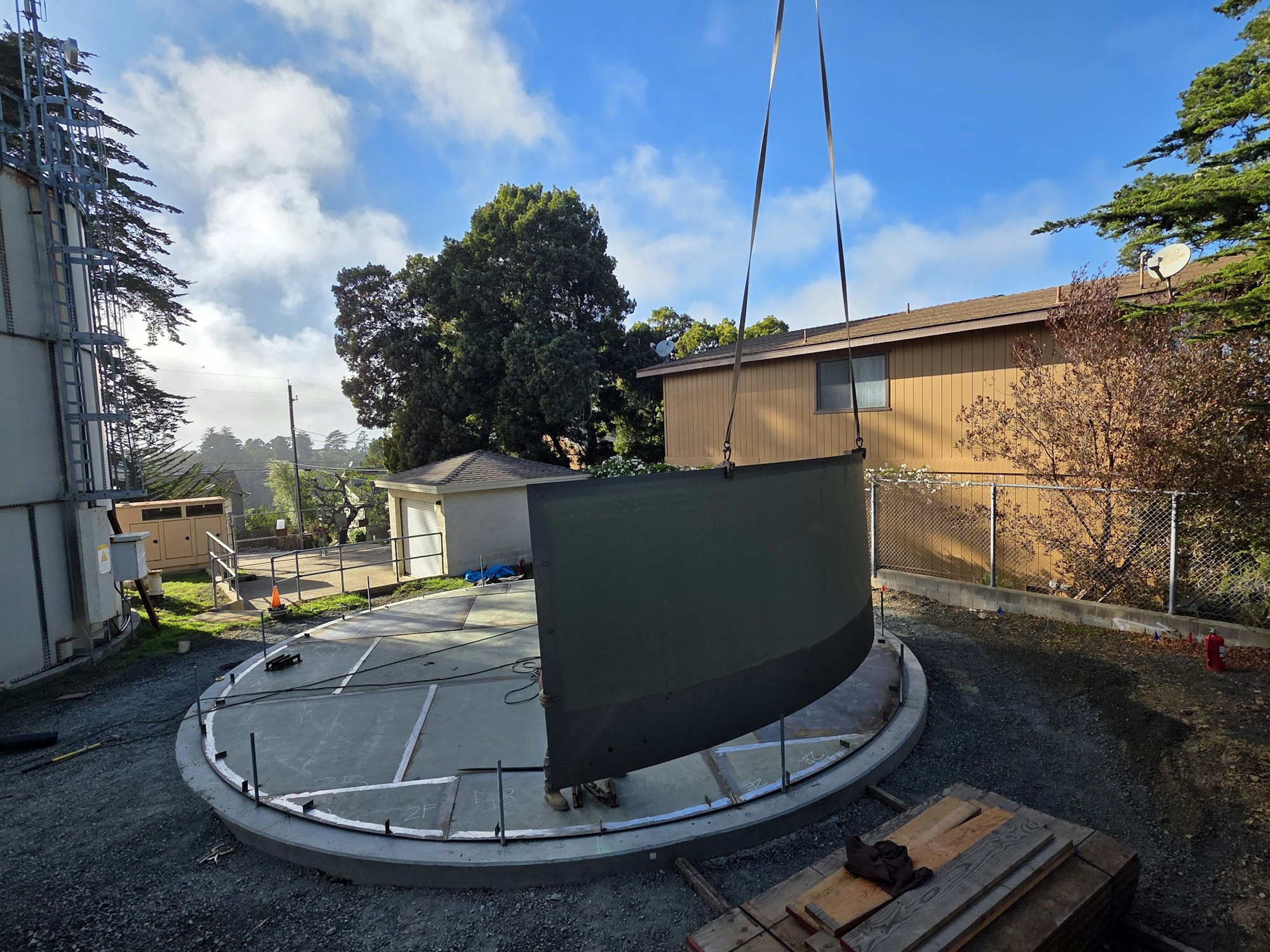 Large metal structure being lifted onto a circular foundation outdoors, surrounded by trees and buildings.