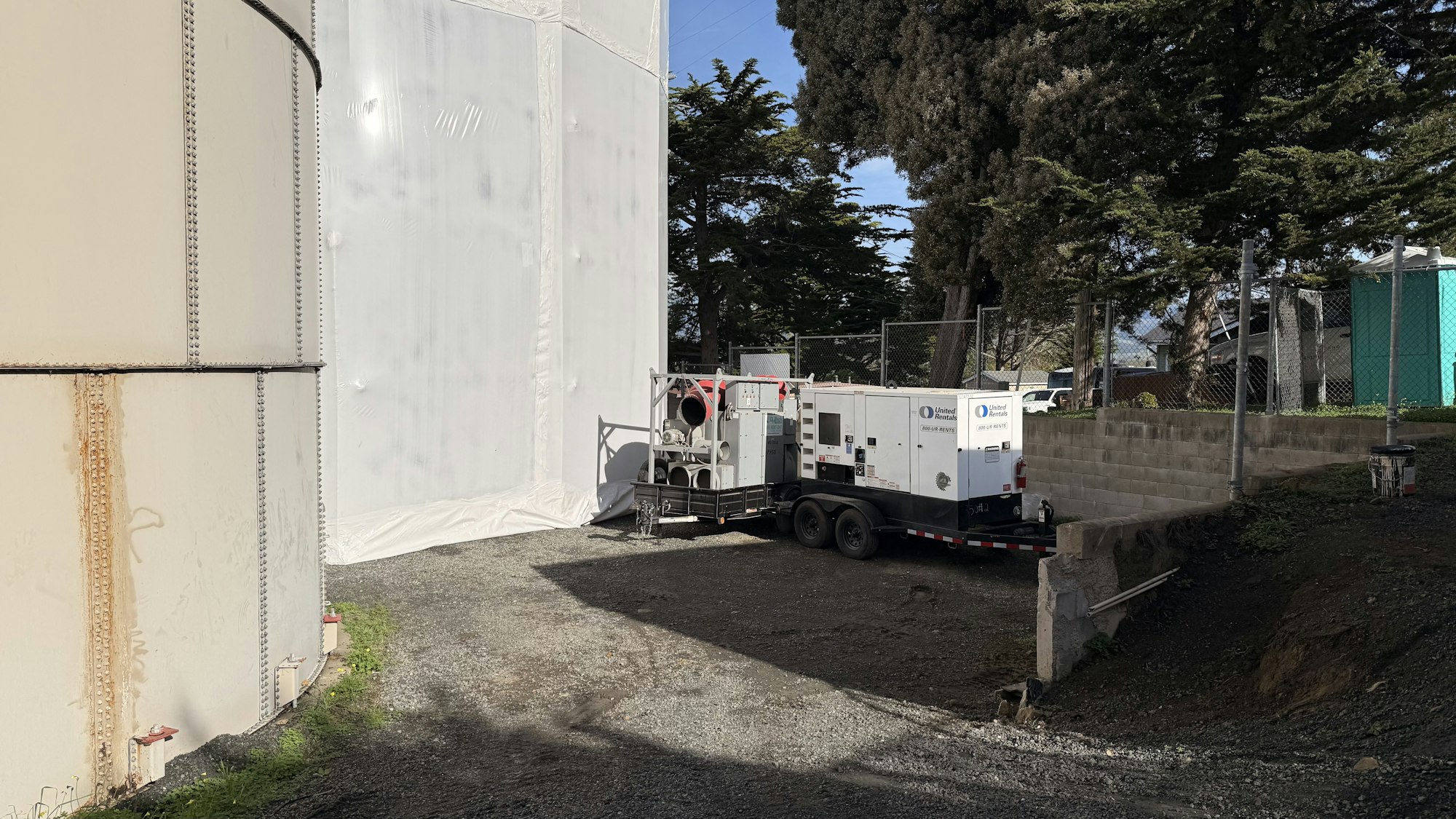 The image shows a utility trailer with equipment parked next to a large tank, surrounded by trees and fencing.