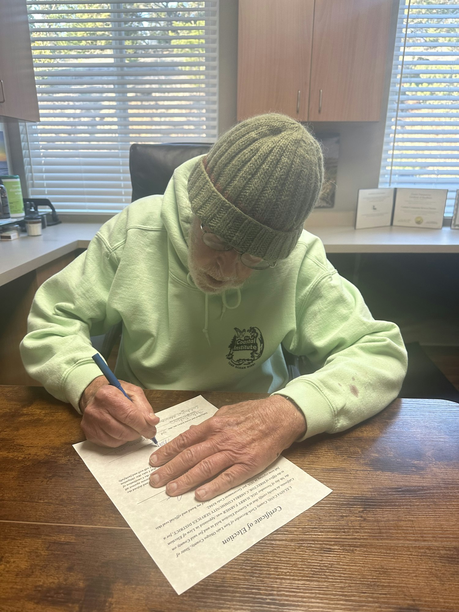 Person in a green hoodie and knit hat signing a document titled "Certificate of Election" at a wooden table.
