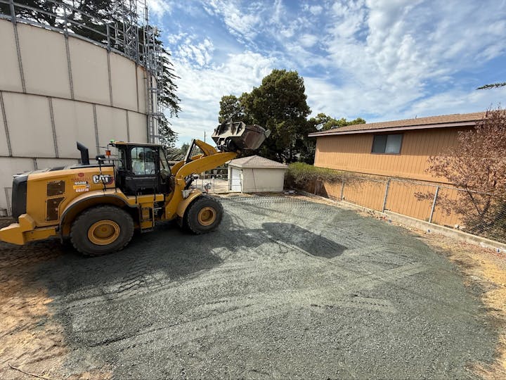 A yellow construction vehicle on a gravel surface near a large storage tank and a house, with a fence and trees in the background.