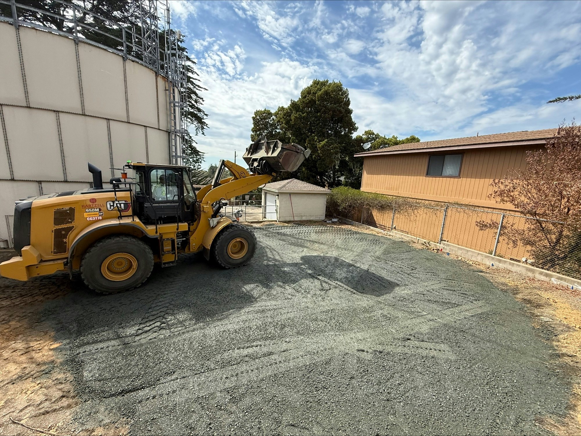 A yellow construction vehicle on a gravel surface near a large storage tank and a house, with a fence and trees in the background.