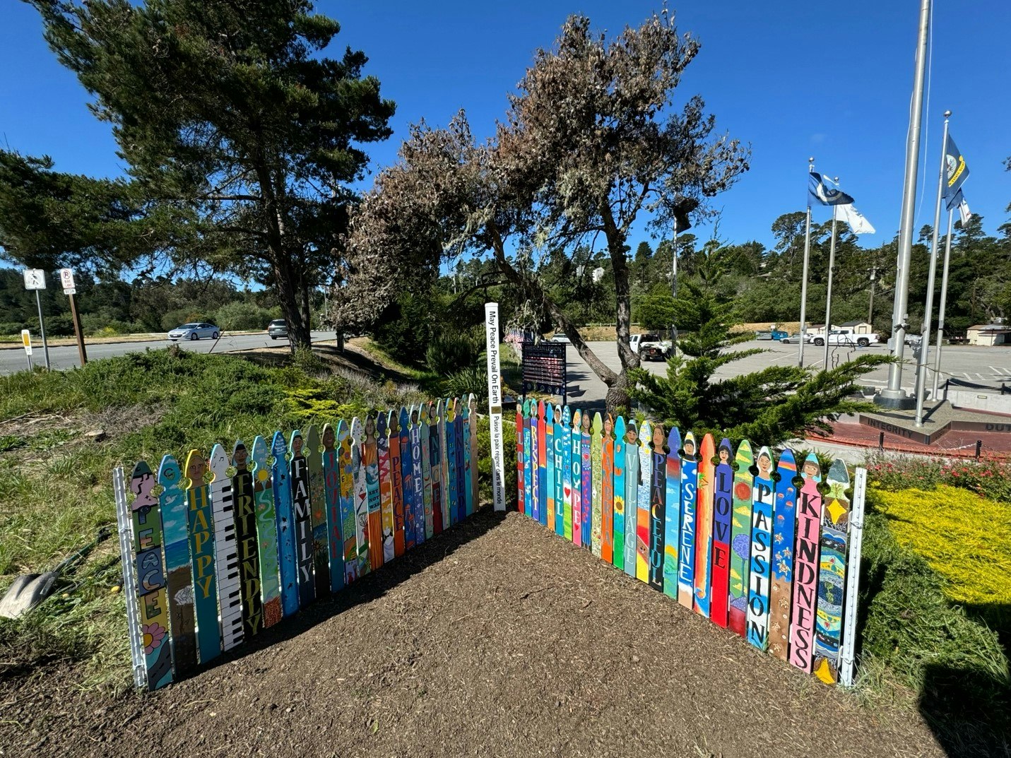 Colorful painted fence with positive words, trees, flags, and a road in the background.