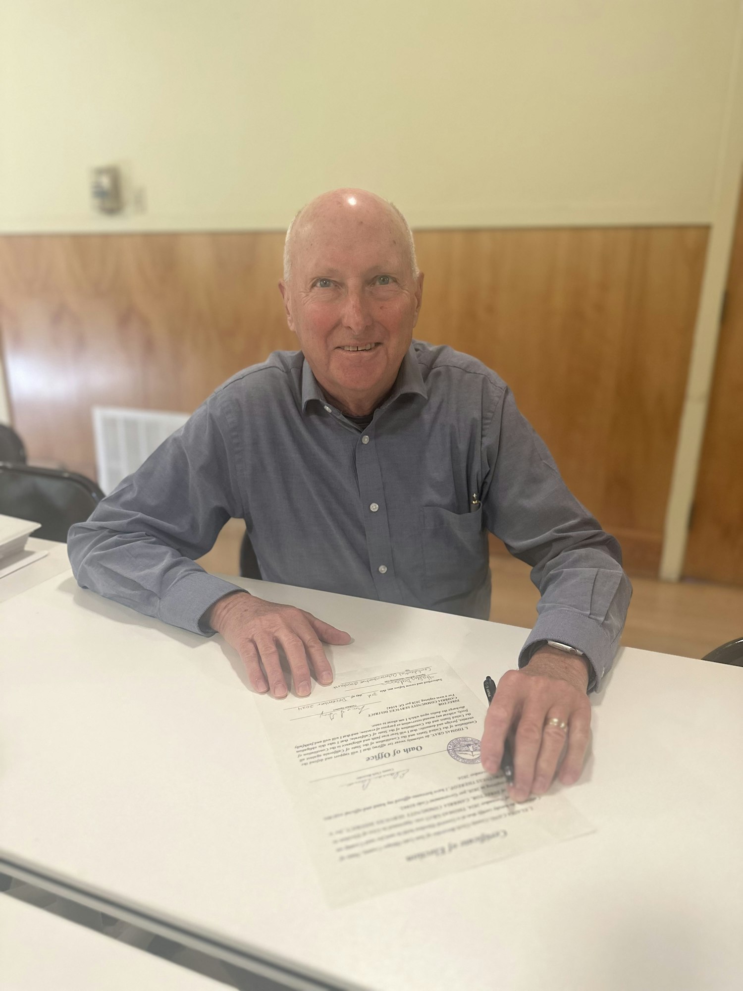 An older man in a gray shirt signing a document at a table.