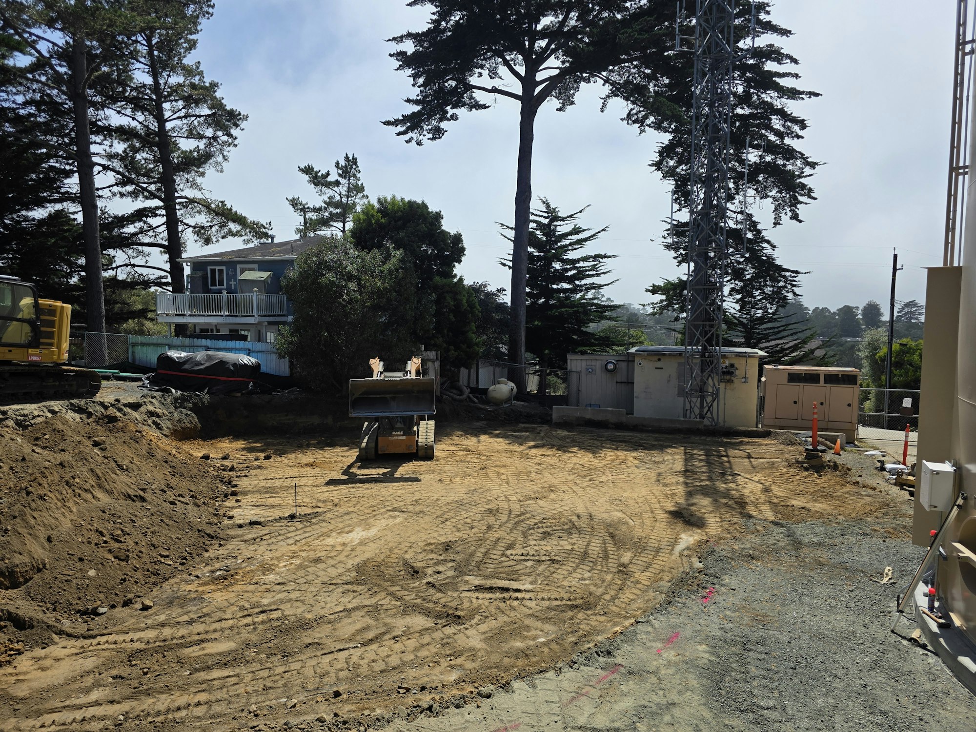 A construction site with heavy machinery, a telecommunications tower, and a house amidst trees in the background.
