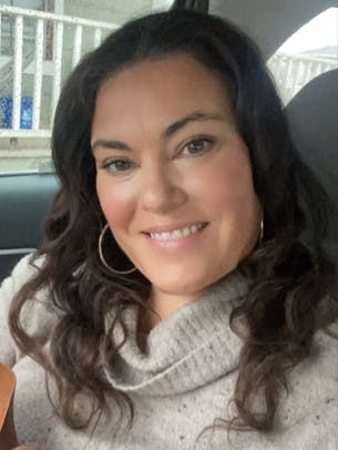 A person smiling in a car, wearing a gray sweater with curly hair and hoop earrings.