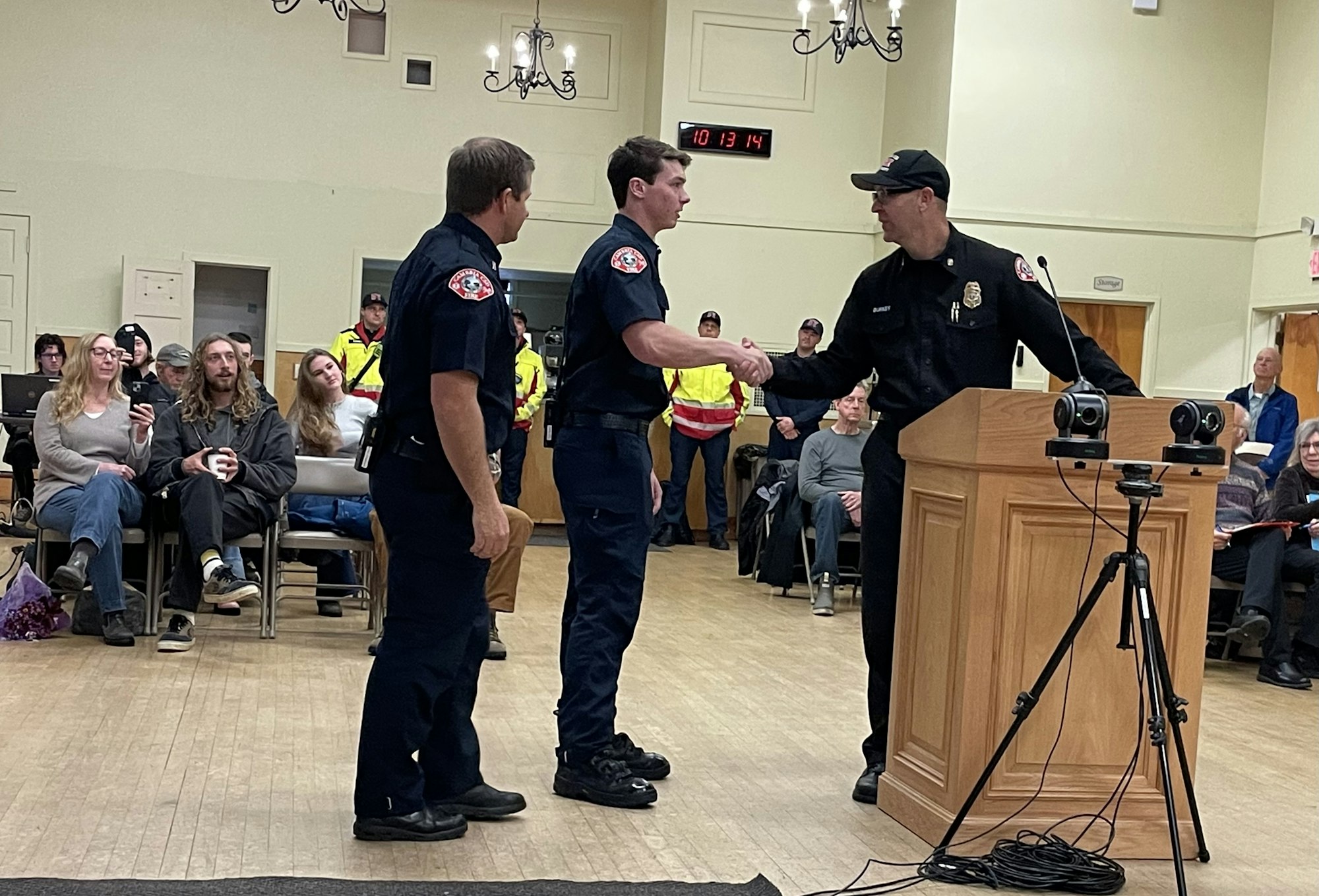 Three uniformed officers at a podium shake hands; seated audience watches.