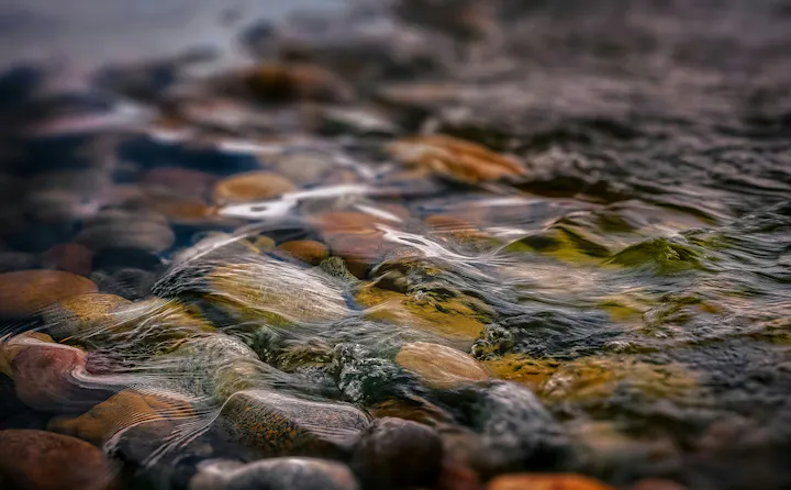 The image shows smooth, colorful pebbles partially submerged in clear water, with gentle ripples reflecting light.