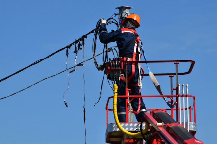 An electrician in safety gear works on power lines from an elevated platform against a clear sky.