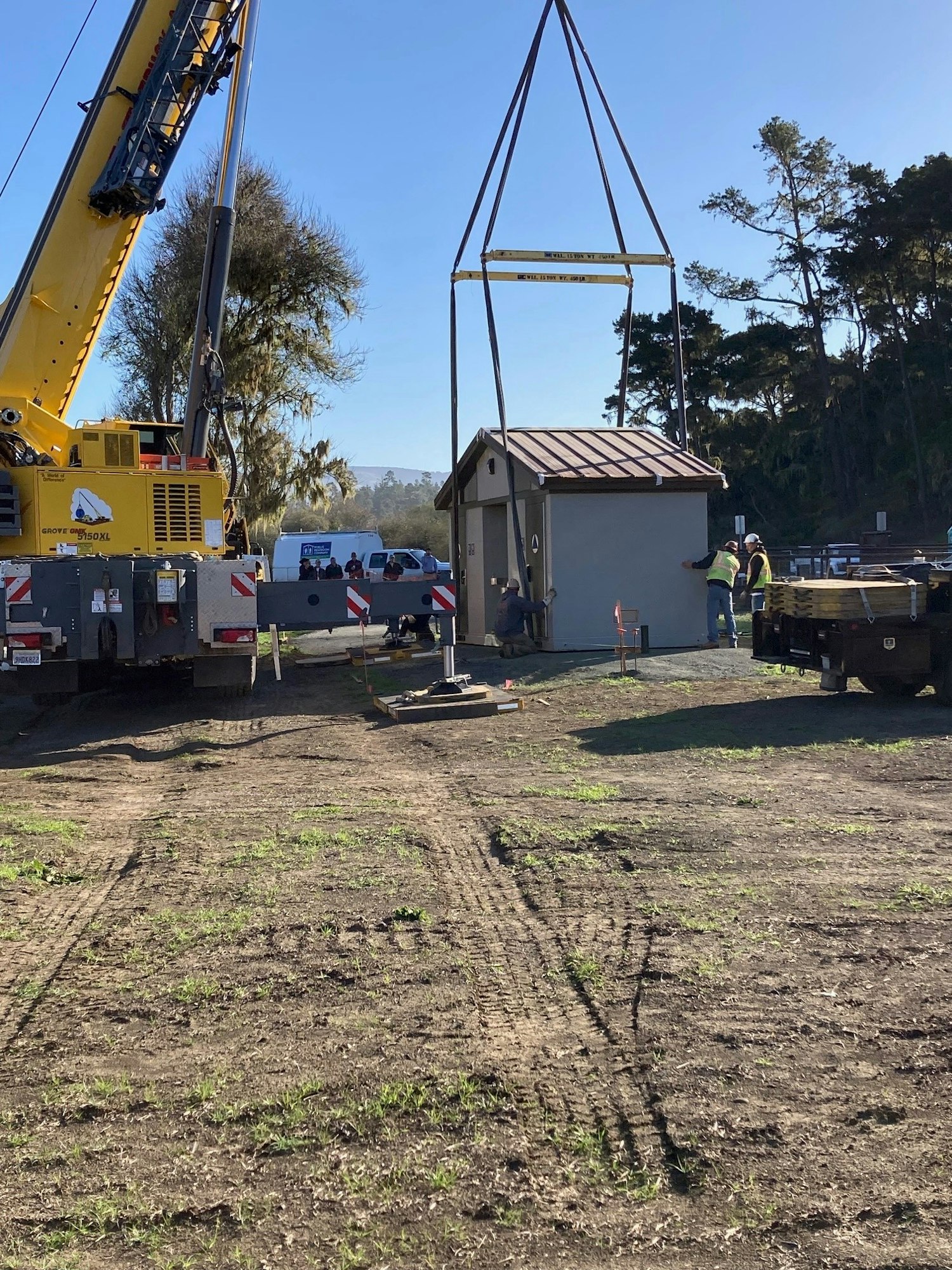 A crane is lifting a small building while workers supervise at a construction site with vehicles and trees in the background.