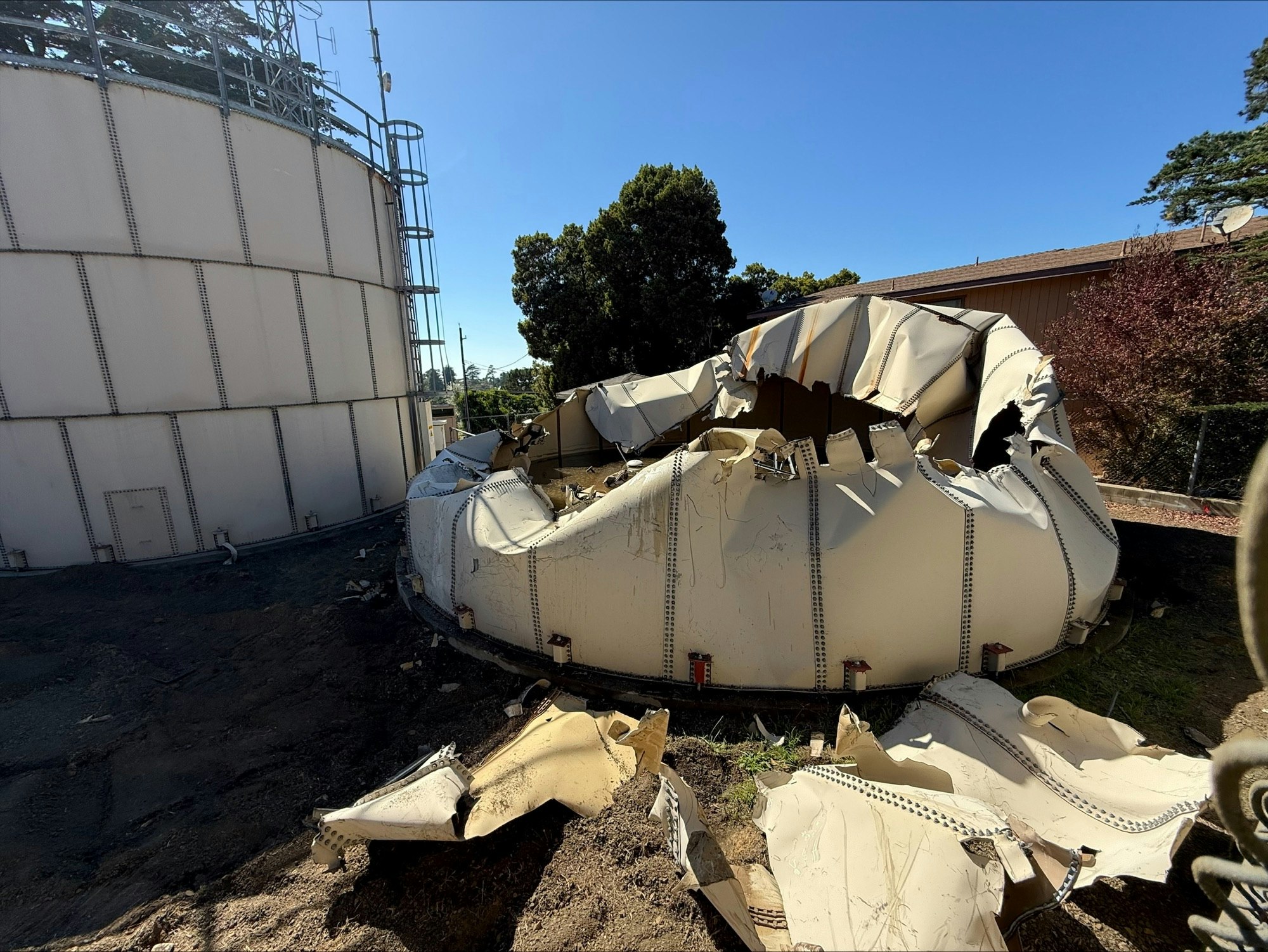 A large, collapsed and mangled metal structure next to an intact metal tank. Trees and a building in the background.